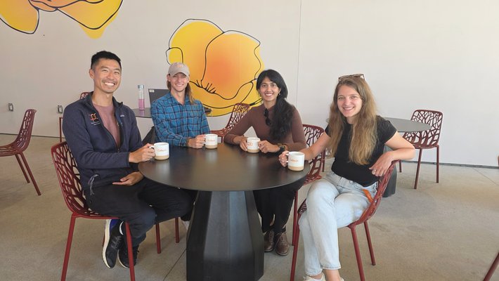 Four postdocs sit at a table with their free mugs and smile at the camera.