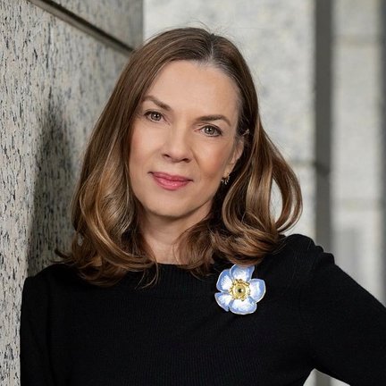 A woman wearing a black shirt with a flower pin leans against a wall and smiles.