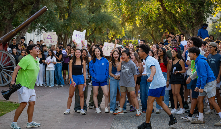 A large group of Caltech students gather outdoors, cheering and holding signs that say "Avery." Two students in the front appear to be leading chants, creating a lively and energetic atmosphere.