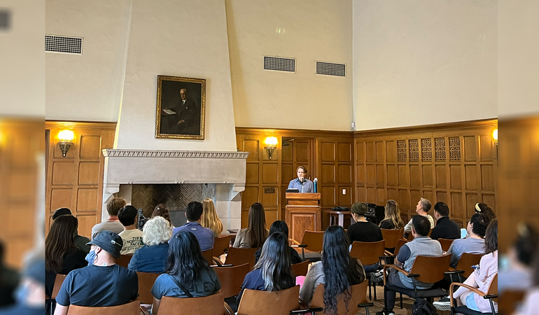 A speaker stands at a wooden podium addressing a seated audience.