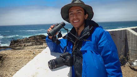 Isaac smiling and holding a pair of binoculars in front of a coastal background