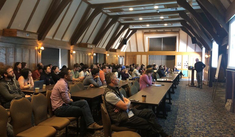 Professor Colin Camerer standing at a lectern giving a talk to an audience of faculty, postdocs and graduate students, seated behind rows of tables at the Chen Institute 2019 Retreat.