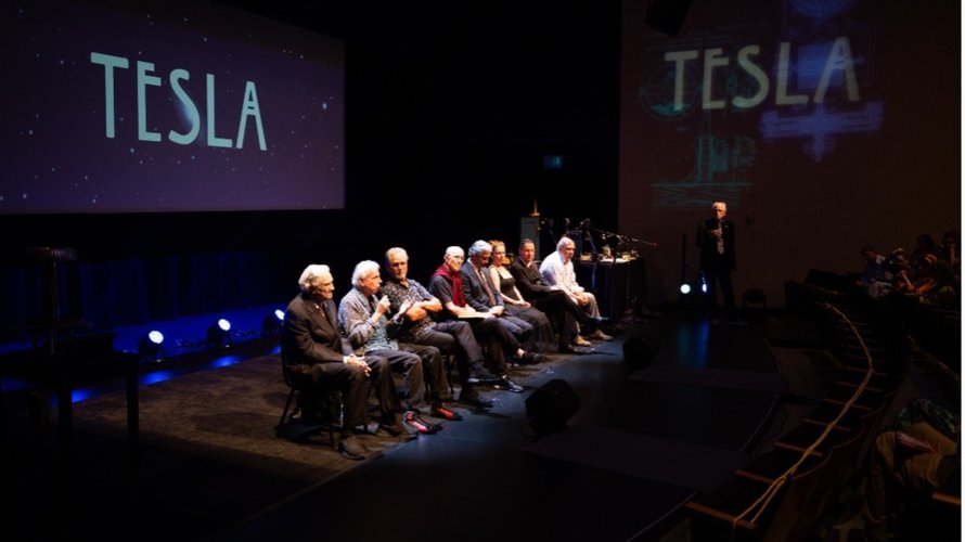 performers sitting on stage with "TESLA" projected on screens