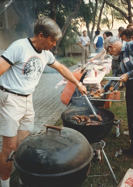 A man in T-shirt and shorts moves meat with a pair of tongs on a smoking charcoal barbecue.