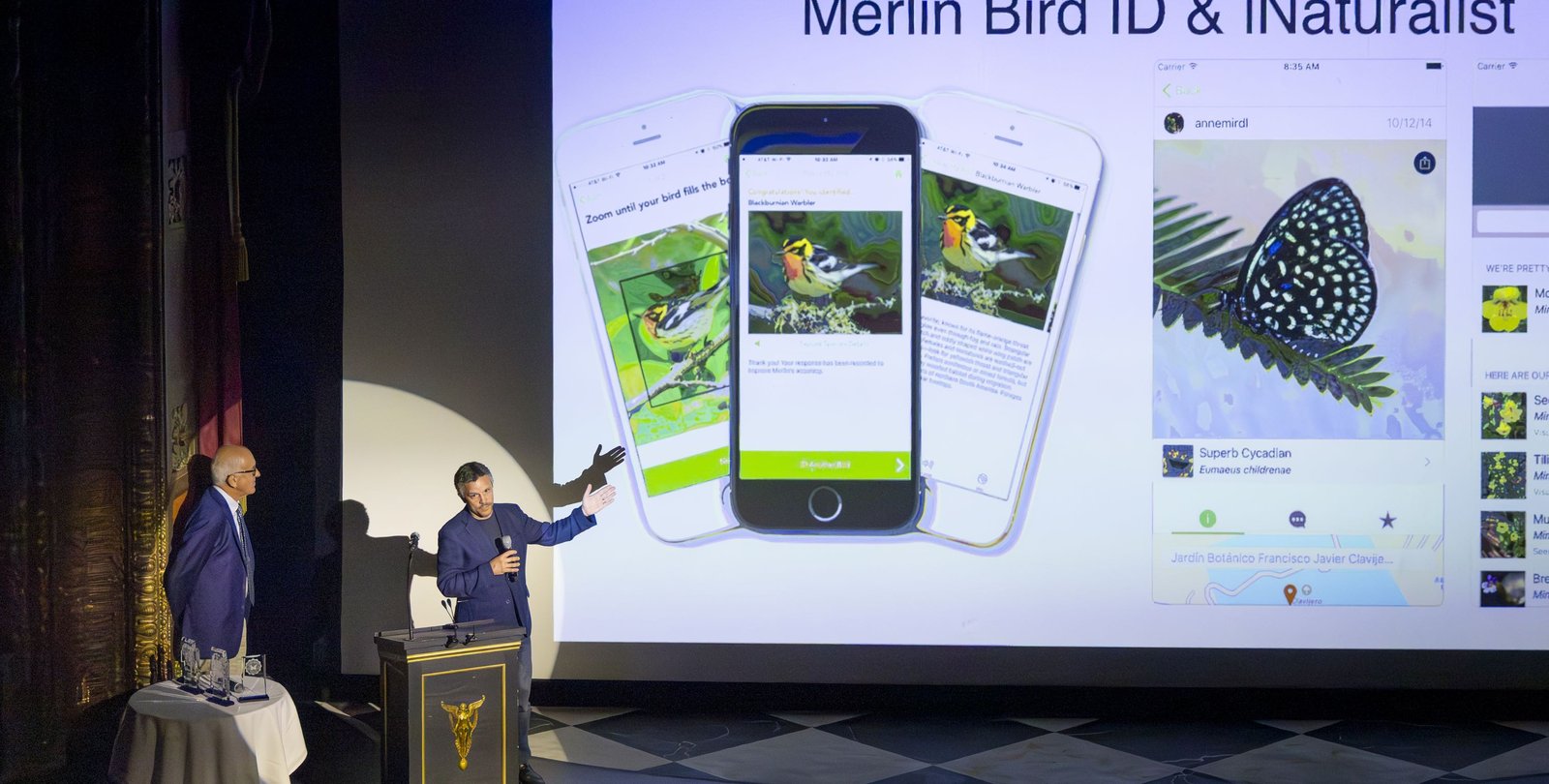 two men on left are presenting on a stage. A slide related to Merlin Bird ID & iNaturalist is shown on a large screen.