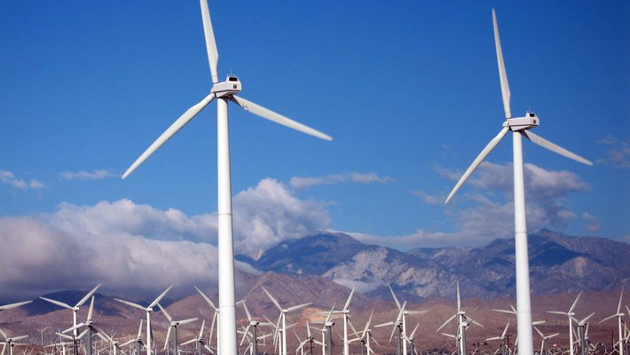 Wind turbines spin in front of a backdrop of clouds and mountains.