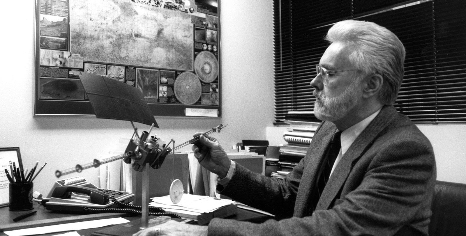 Arden Albee sits at a desk wearing a tie and sport jacket, looking at a model spacecraft for a Mars mission. There is a large poster on the wall titled "An Explorer's Guide to Mars."