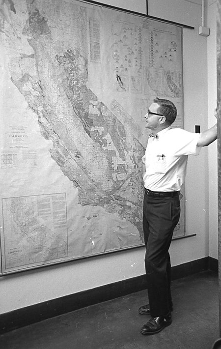 Arden Albee stands, in a white short-sleeved shirt and bow tie, looking at a very large relief map of the state of California.