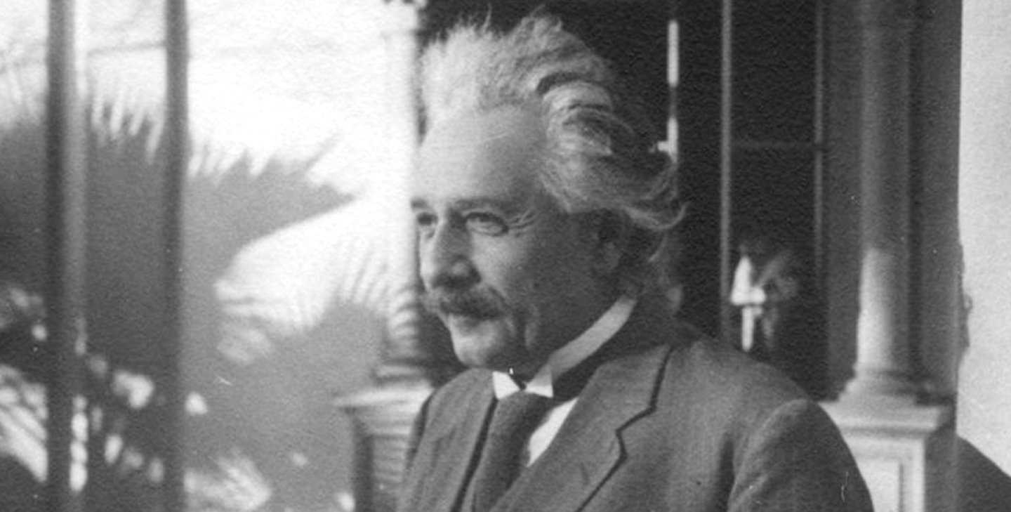 A black and white photograph of Albert Einstein. He has silver hair brushed back from his face and a dark mustache. He wears a three-piece suit and tie.