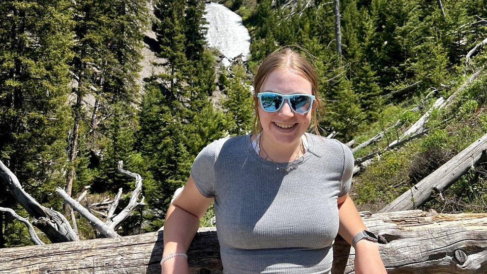 A person leans on a log in front of an evergreen forest.