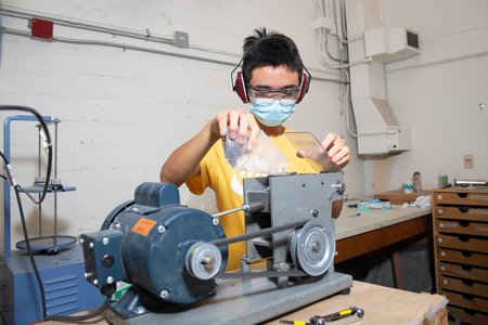 A person in safety glasses and a KN95-style mask feeds rocks from a plastic bag into a crushing machine in a laboratory.