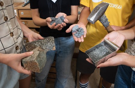 The hands of four people hold rocks in different stages of crushing: whole, sawn into fragments and cleaned and sanded, broken with a sledgehammer (also shown), and crushed in the rock-crusher's tray,