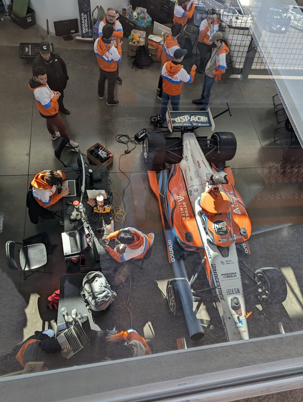 an overhead view of Caltech's autonomous race car and about a dozen members of the team that support it wearing orange, blue, and white Caltech jackets