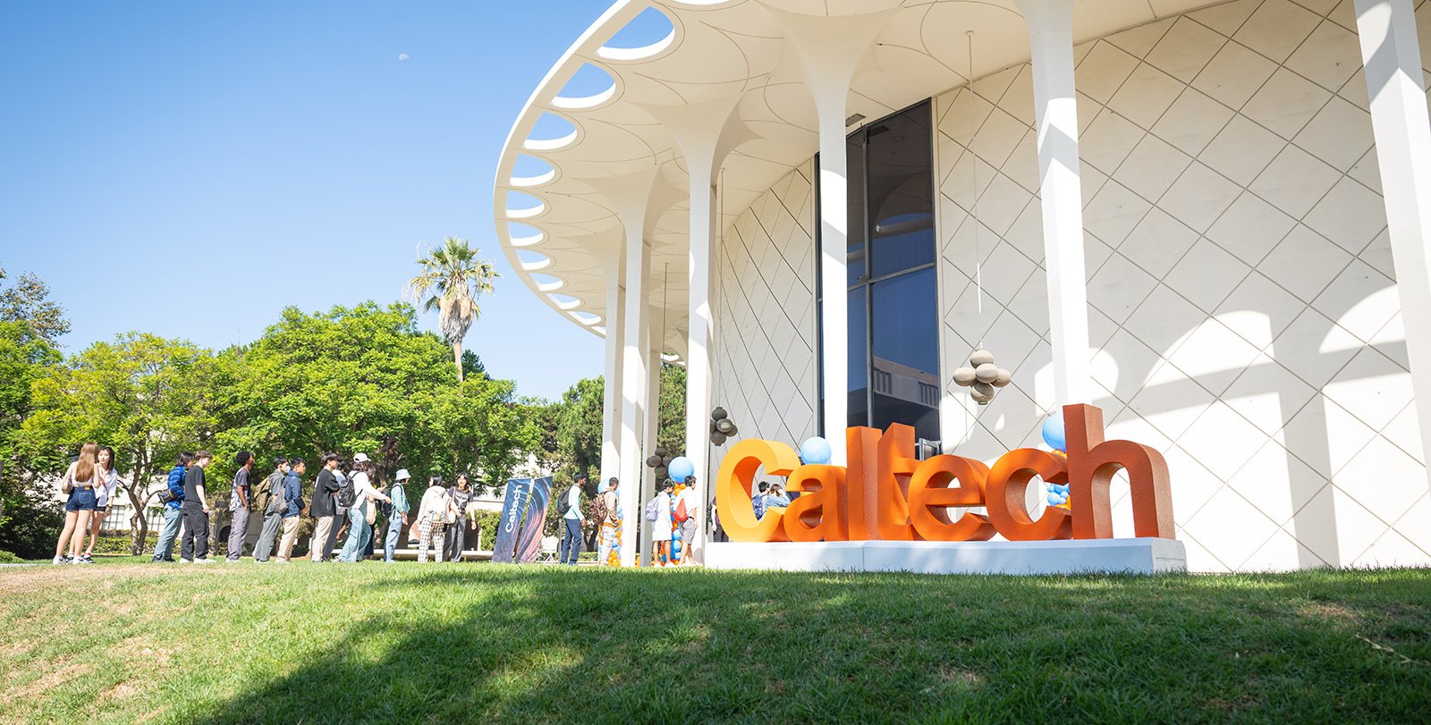 Students walk into the Beckman Auditorium. In the foreground are large orange letters spelling out "Caltech."