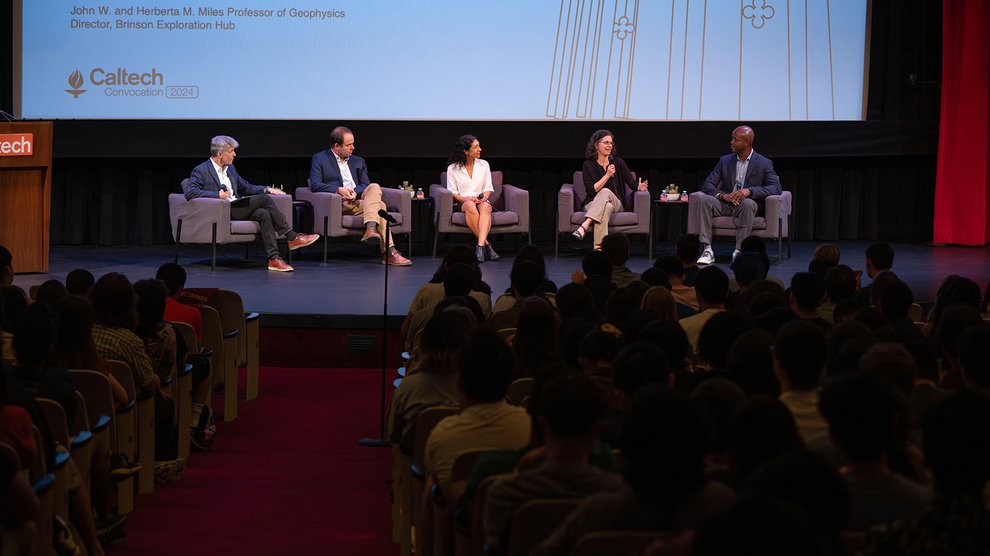 Five people sit in armchairs on a stage in front of a crowd.