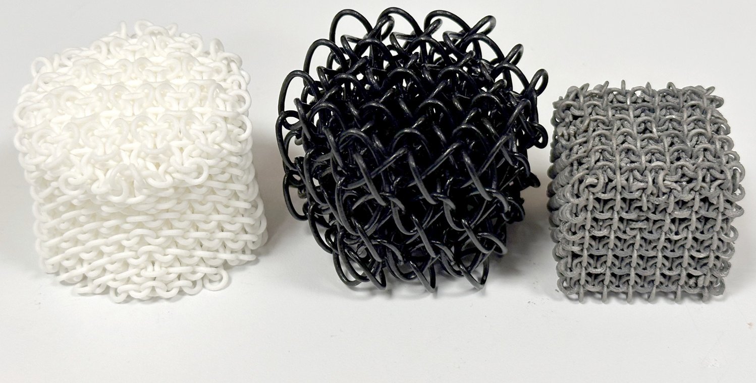Three polycatenated architected materials printed out in three different materials: nylon, TPU, and steel. The nylon PAM is white and cylindrical; the TPU one is black and roughly spherical; the steel one is dark gray and cubical.