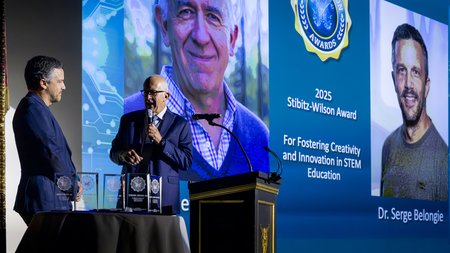 Pietro Perona speaking with a microphone in front of a large screen showing details of the award he and Serge Belongie received at the pictured event. Belongie is also in the shot along with the awards themselves.