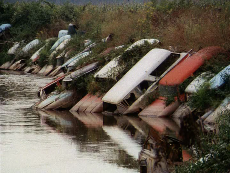 A still frame from archival footage. The frame shows rusting cars on the bank of a river, partially resting in the water.