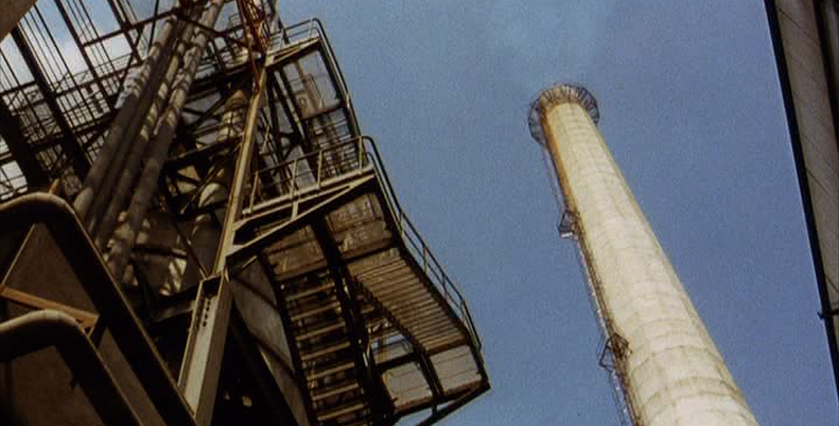 A grainy still from a vintage film. The camera points upward through the infrastructure of an oil refinery, showing a smokestack set against a blue sky.