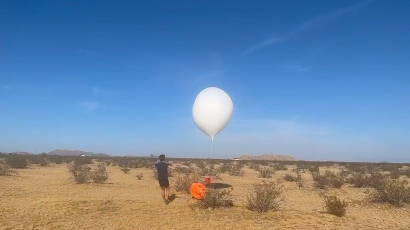 a large white balloon tethered to an orange payload has been released by a person wearing black in a desert landscape with a bright blue sky