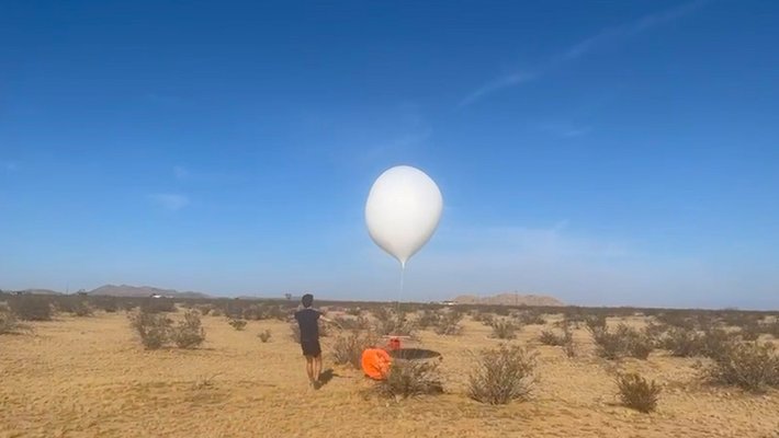 a large white balloon tethered to an orange payload has been released by a person wearing black in a desert landscape with a bright blue sky