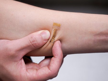 A smart bandage is shown adhered to the skin of a forearm. Two fingers of another hand pinch the skin on either side of the bandage, showing how it stays on.