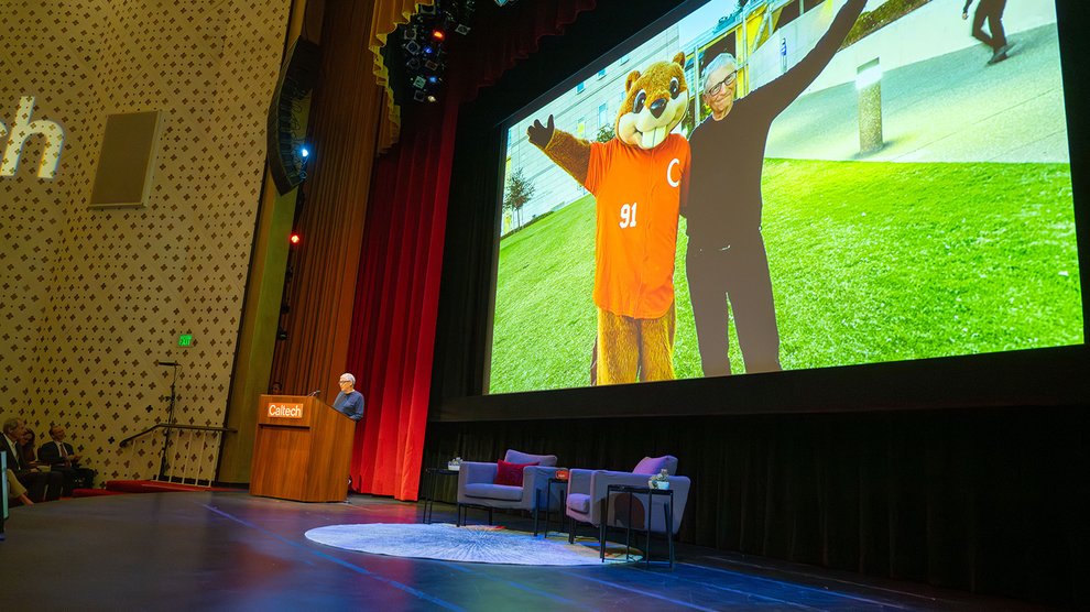 A screen projection showcases a photo of Bill Gates posing with Caltech's mascot while Bill Gates speaks on stage