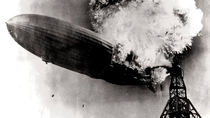 A large zeppelin, partially consumed by flames, begins sinking toward the ground next to its mooring mast. The image is a black and white photograph.