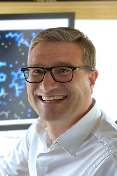 A white man wearing a white button-up shirt and dark-rimmed glasses smiles at the camera. A computer screen with chemical structures can be seen in the background.
