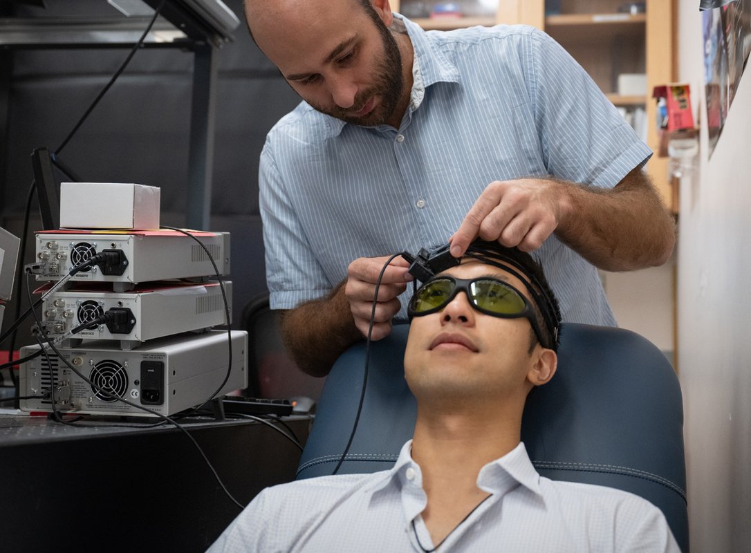 One man is putting a headset-based device on another man who is sitting in a chair in front of him. The man in the chair is wearing sunglasses