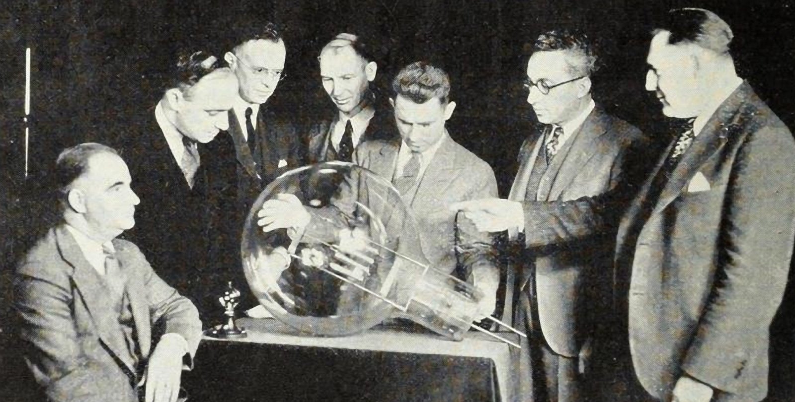 A black and white photograph from 1929 of seven men in suits, one seated and six standing, looking at an electric lightbulb the size of a beach ball resting on a table before them.