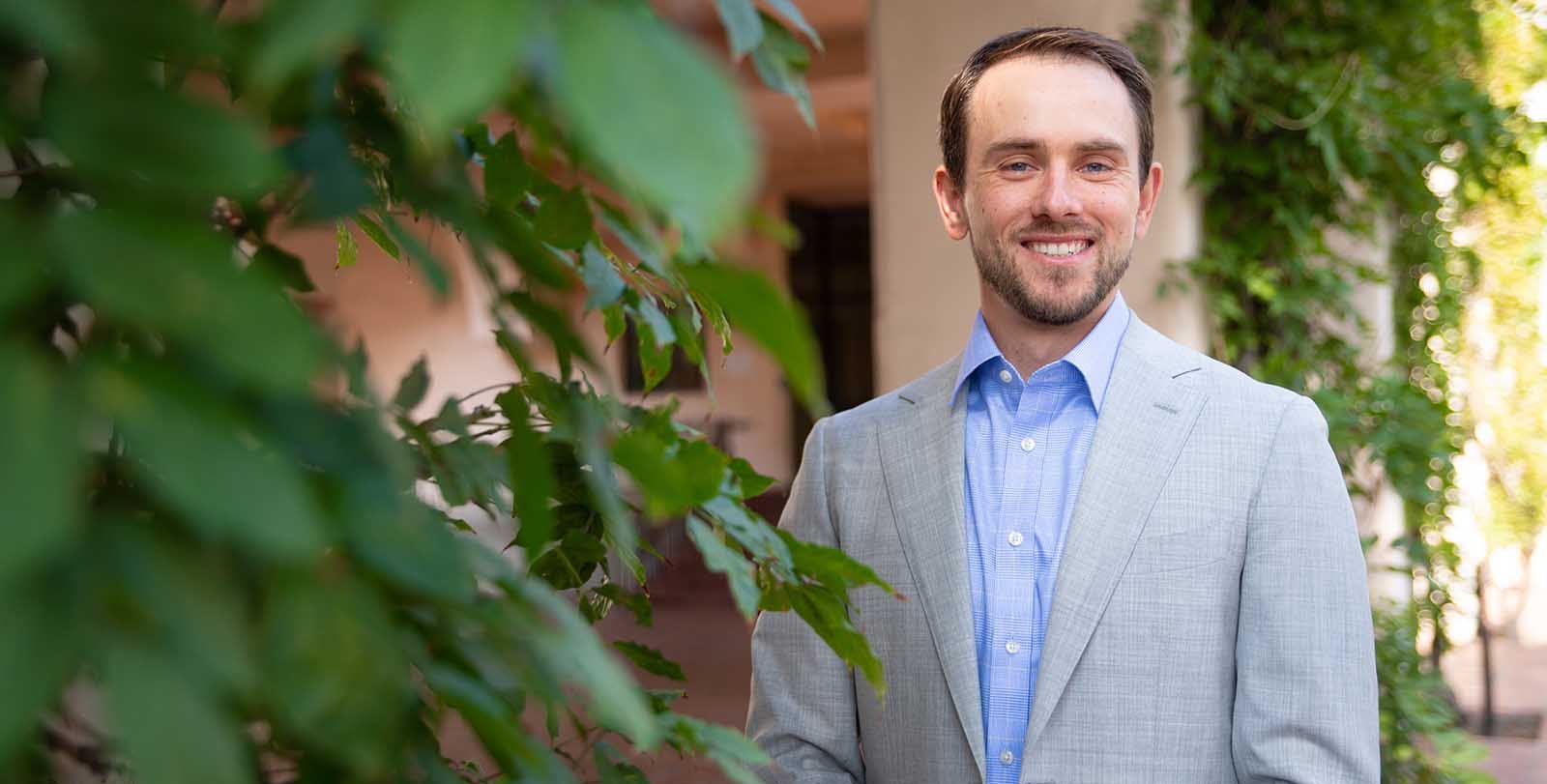 A portrait of Pawel Janas. He stands outside smiling and wearing a suit. There are columns with vines behind him.