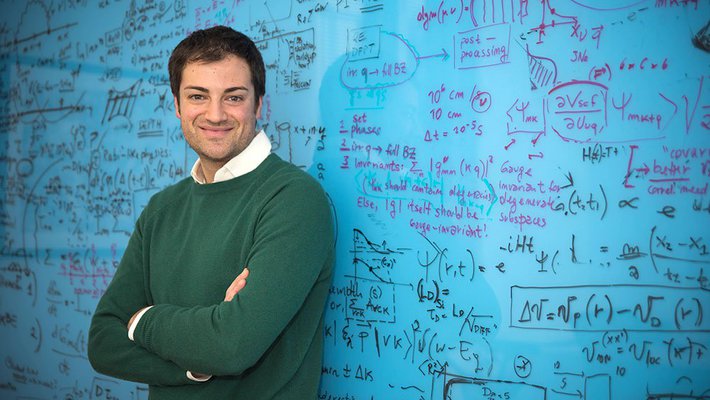 A portrait of Marco Bernardi. He stands in front of a blue chalkboard covered in writing with his arms crossed.