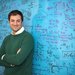 A portrait of Marco Bernardi. He stands in front of a blue chalkboard covered in writing with his arms crossed.