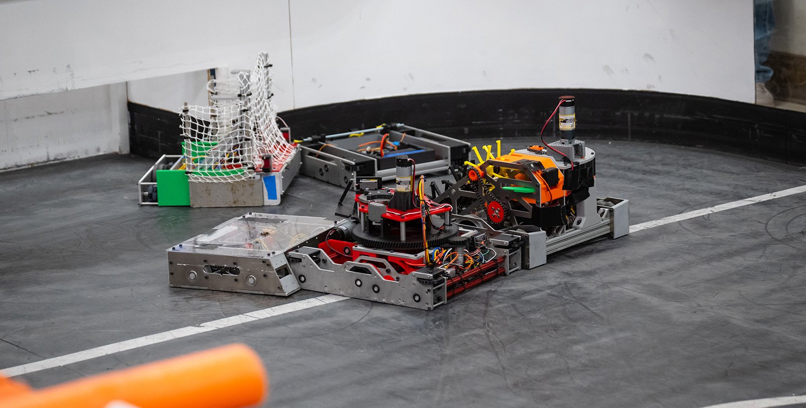 A close-up photo showing five robots battling each other, almost tangled up, on a custom-built hockey rink with white walls, an orange rim, and a black ferromagentic surface.