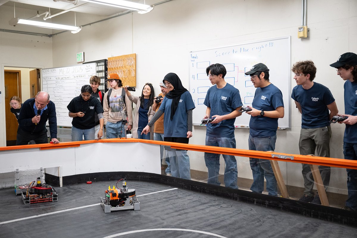 Five students wearing dark blue Caltech MCE (mechanical engineering) shirts control and watch from the sidelines of a custom-built bot hockey rink in a subbasement machine shop. Instructor Mello and others watch the match with interest.