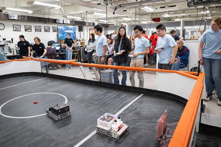 Students in light blue t-shirts look into the rink during the ME72 competition. The mechanical engineering machine shop as well as other students and spectators are visible in the background.