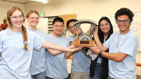 Six students in light blue t-shirts smile and hold up a gear-shaped trophy.