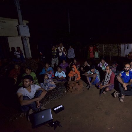 Many people sit on the ground in a meeting, looking toward a projection. Otherwise, everything is dark.