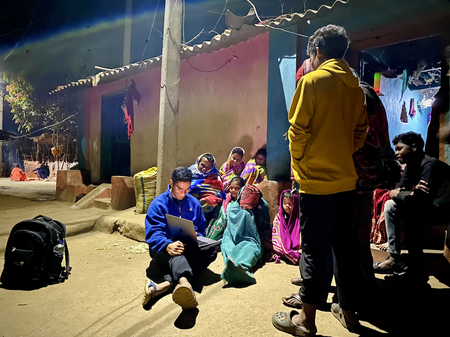 Several children and others sit and stand around Eshaan Patheria who has a laptop on his lap. The scene is illuminated by a street lamp, and a lightbulb is on in the house behind them.