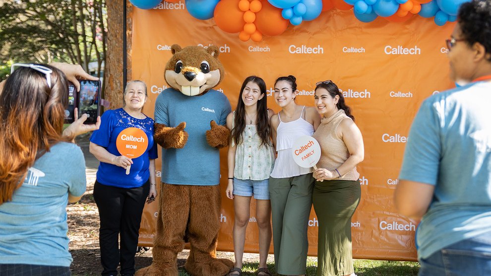 A family poses for a photo with a Berni the beaver mascot in front of an orange Caltech backdrop.