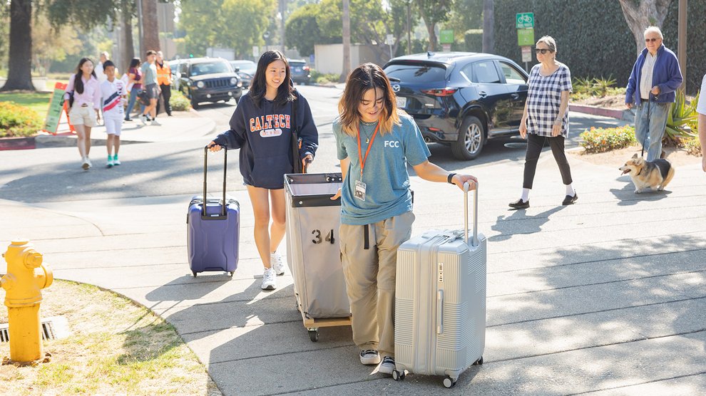 Two students walk on campus, pulling suitcases with them.