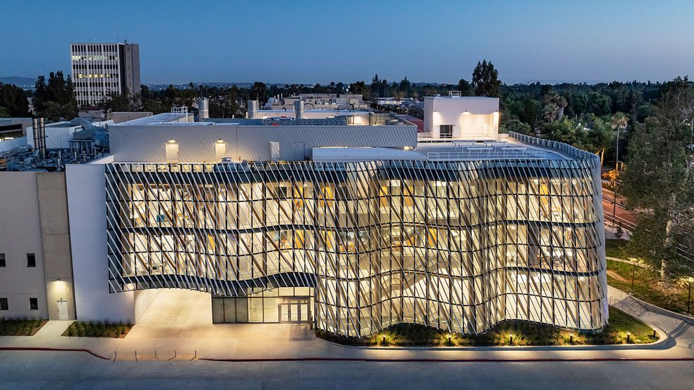 A building with a glass and metal wall with lights on inside that are visible at dusk.