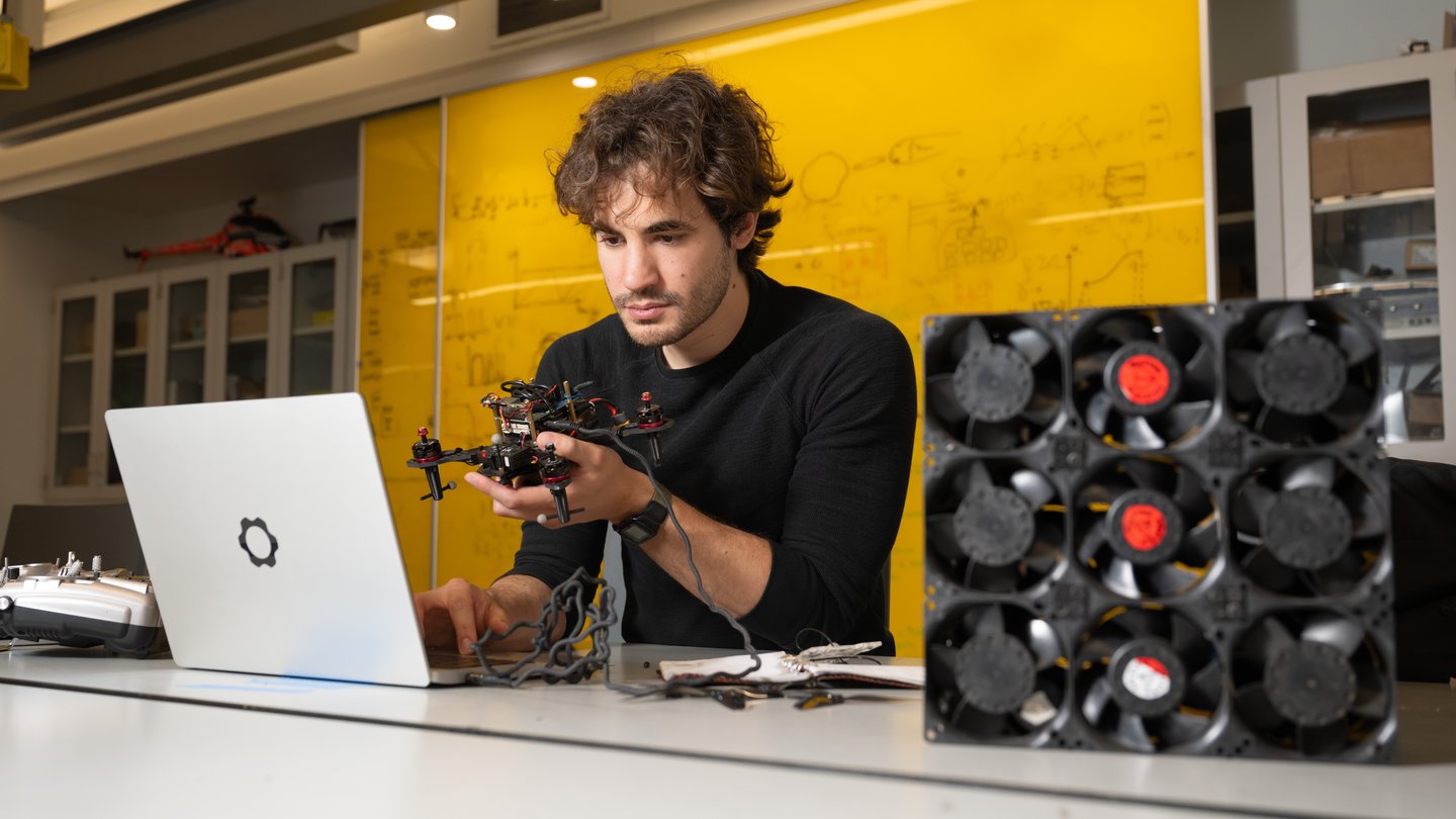 Alejandro Stefan-Zavala sits in front of a laptop with a drone in hand and small fans off to his side.