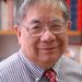 Sunney Chan wears a shirt and tie and smiles at the camera in this portrait. Bookshelves can be seen in the background.