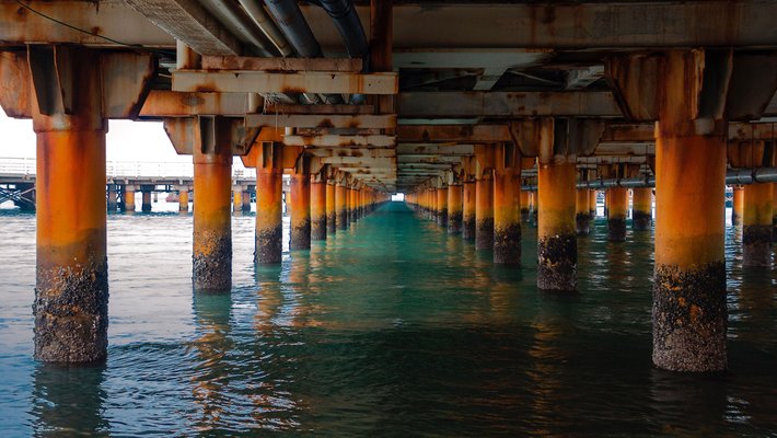 An image of the underside of a rusty bridge over a body of water.