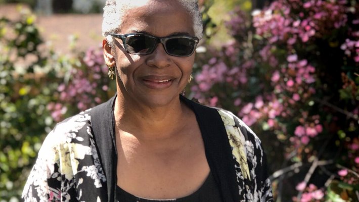 Woman in sunglasses and floral jacket standing outside in front of flowers