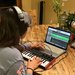 A young woman, her back to the camera, is wearing headphones and working on a computer and MIDI board.