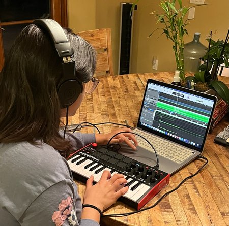 A young woman, her back to the camera, is wearing headphones and working on a computer and MIDI board.