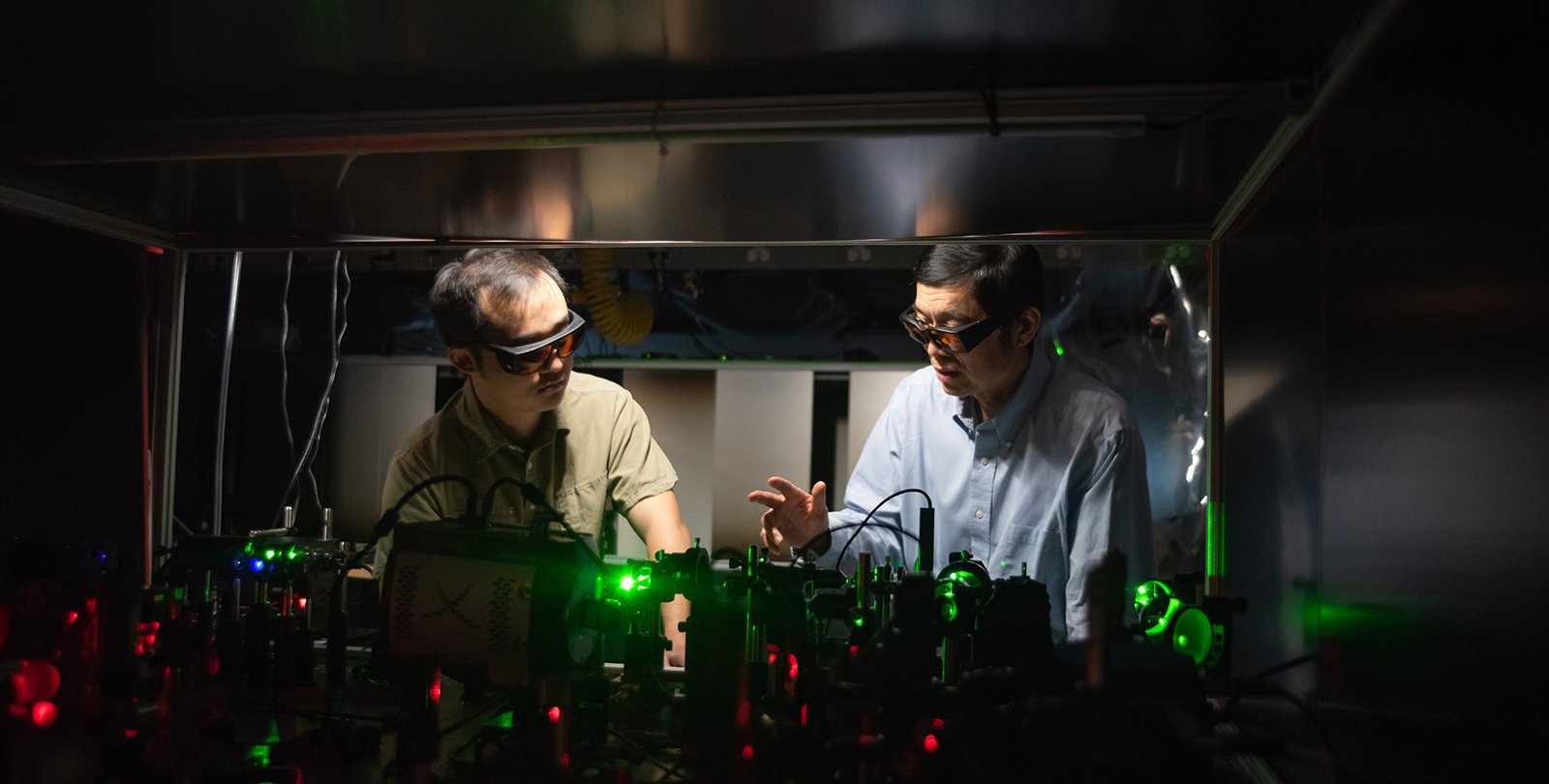 Two men wear safety glasses and look at an optical apparatus consisting of many lenses and mirrors and prisms in the foreground.